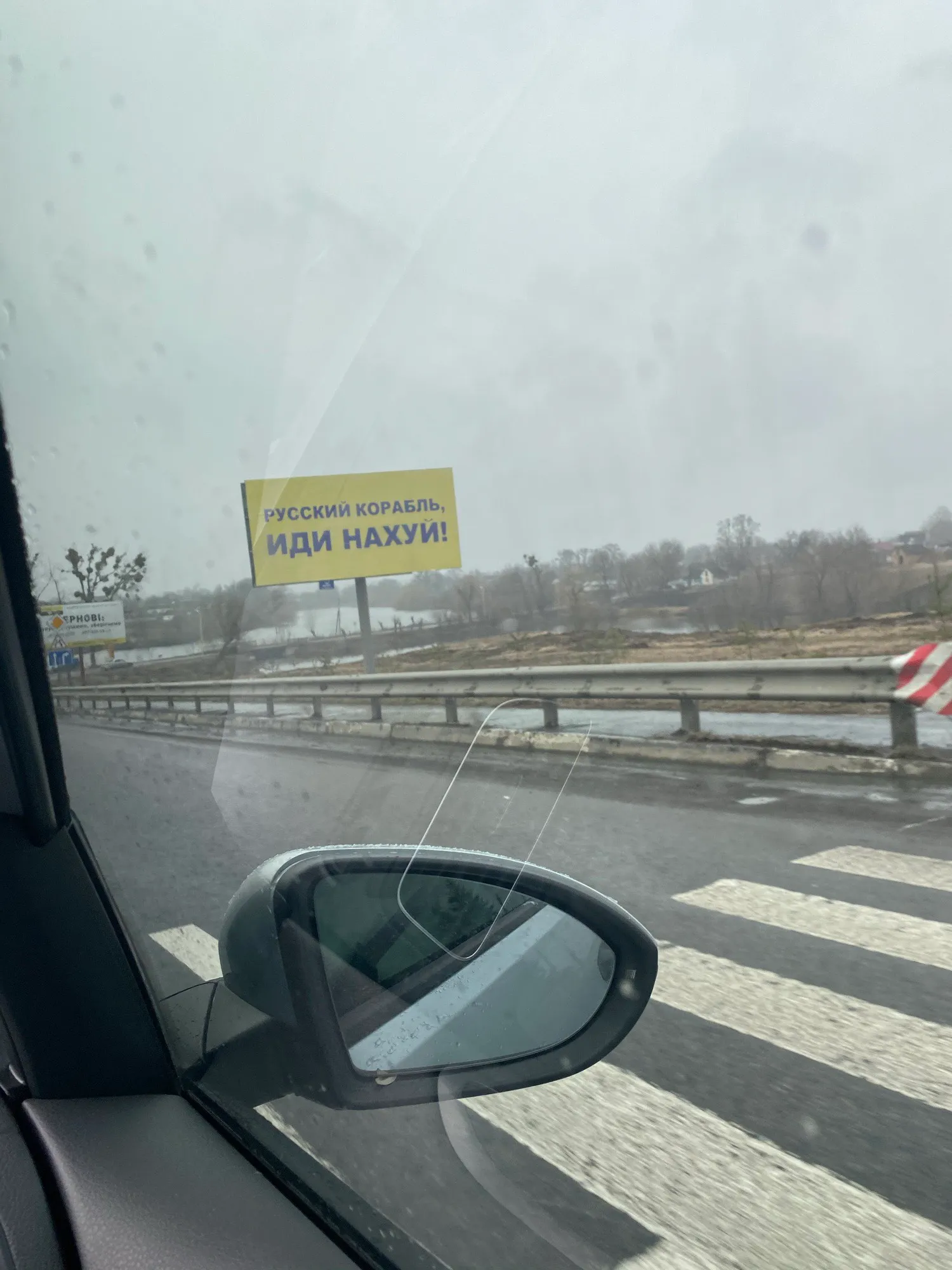 A yellow Ukrainian road sign reading “Russian warship, go f*** yourself”, photographed through a car windscreen on a wet grey day